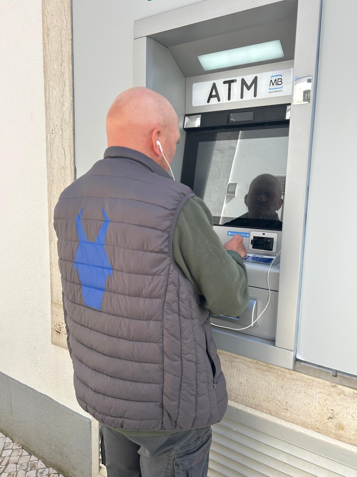 Um homem careca, de pele clara, com colete acolchoado cinzento (com o símbolo da Associação Cabra Cega azul grande nas costas), camisola verde e calças escuras, está de costas a usar uma caixa Multibanco instalada numa parede exterior. Tem um auricular com fio branco no ouvido esquerdo.  Em cima lê-se “ATM” e aparece o logótipo “MB Multibanco”. Há uma luz retangular por cima do ecrã. O ecrã está escuro e reflete a cara do homem. 